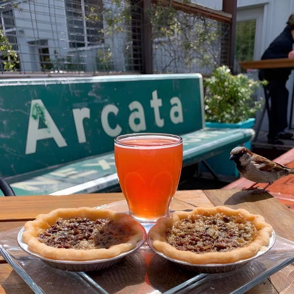 Two pecan pies and a glass of orange drink on a wooden table with an "Arcata" sign.