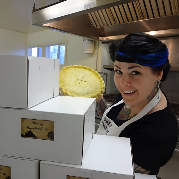 A smiling baker holds a freshly baked pie next to stacked Slice of Humboldt Pie boxes.