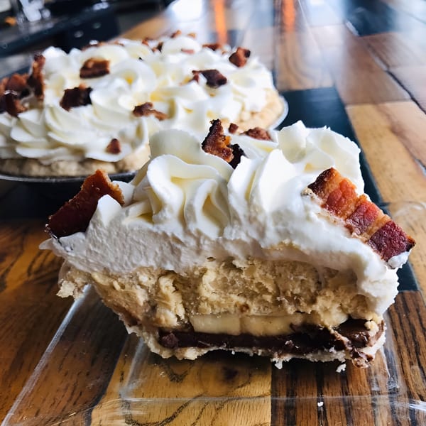 A slice of banana cream pie with whipped cream and bacon bits on a wooden table.