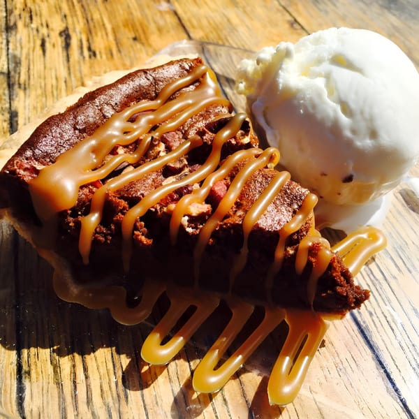 A slice of chocolate pecan pie drizzled with caramel, served with a scoop of vanilla ice cream.